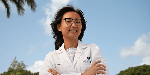 Student in a white coat crossing her arms with a palm tree behind her